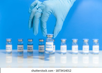 A Hand Of Scientist, Doctor, Nurse, Medic, Intern In Latex Gloves Holds Covid Coronavirus Vaccine Vial, A Lot Of Bottles On The Background. Isolated On Blue. Concept Of Confrontation With Viral Threat