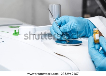 Hand of scientist using a stainless spatula to scoop sample solid from vial to the Fourier Transform Infrared Spectroscopy FTIR pedestal instrument for substance analysis and chemical characterization