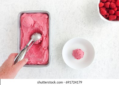 Hand scooping out homemade raspberry ice cream from a metallic tub. Scoop of ice cream in a bowl and fresh raspberries on white marbled background.