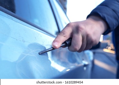 Hand with a screwdriver or a large nail scratching the paint surface on the vehicle door, close-up. The criminal is engaged in vandalism. Revenge neighbor, business partner or offended lover