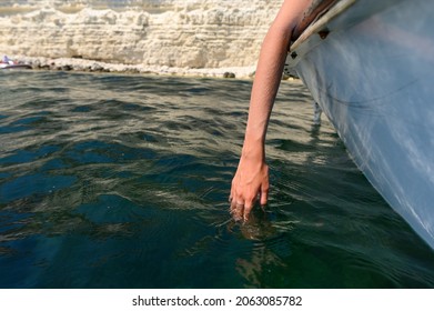 Hand In Sea Water From A Boat On The Background Of Rocks.