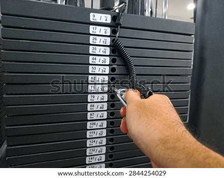 Hand selects weights from organized stack. Fingers grip tool, targeting specific plate. Stack labeled, showing varied weight increments. Dark gray plates, orderly arranged in rows