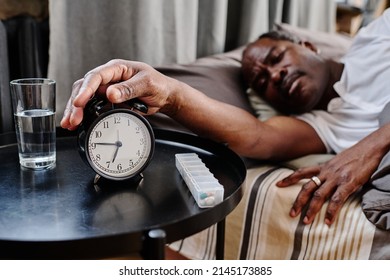 Hand Of Senior African American Sleepy Man Pressing Buzz Bell Of Alarm Clock On Small Round Table In The Morning While Staying In Bed