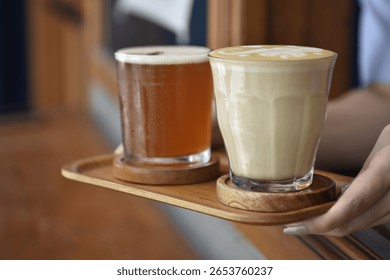 A hand serving two drinks a creamy latte and a cold brew on a wooden tray. Ideal for illustrating café service, hospitality, or beverage lifestyle themes.
