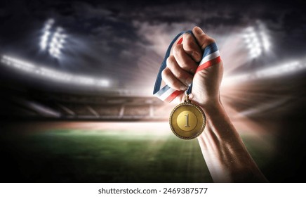 Hand showing gold medal with engraved number 1 and athletics stadium behind at night - Powered by Shutterstock