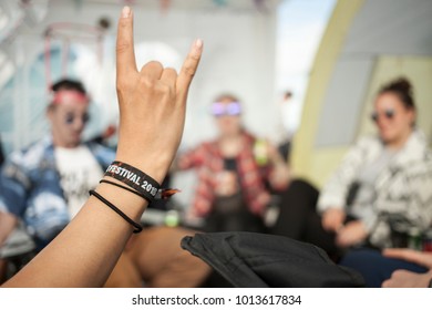 A hand showing the sign of the horns, whichs usually refers to the appreciation of rock music. The wristband says Festival 2018. A group of friends is sitting, chatting and drinking in the background. - Powered by Shutterstock