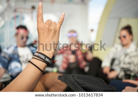 A hand showing the sign of the horns, whichs usually refers to the appreciation of rock music. The wristband says Festival 2018. A group of friends is sitting, chatting and drinking in the background.