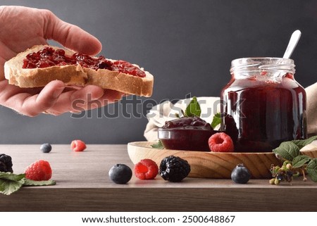 Hand showing slice of bread with berries jam and wooden kitchen bench with containers with jam and breakfast products and isolated dark background. Front view.