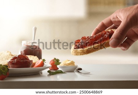 Hand showing slice of bread with strawberry jam and white kitchen bench with containers with jam and breakfast products with kitchen in background. Front view.