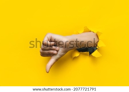 Hand showing a thumb down through ripped hole in bright yellow paper background. Concept of dislike and disapproval gesture.