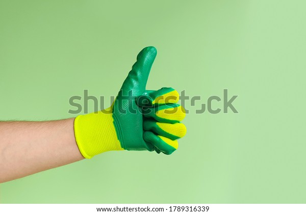 Hand\
sign thumbs up. Male hand in a glove for working in the garden,\
vegetable garden on a green background. Copy space.\

