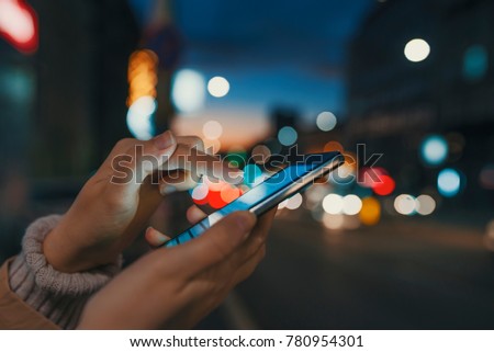 Hand with a smart phone in a dark. Female using her mobile phone, city skyline night light background. Girl pointing finger on screen smart phone on background illumination color light in night city