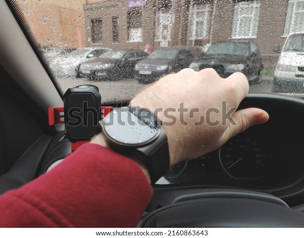 A hand with a smart watch on the\
steering wheel of the car. Reflection of water droplets on the\
glass of the watch and drops on the glass of the\
car