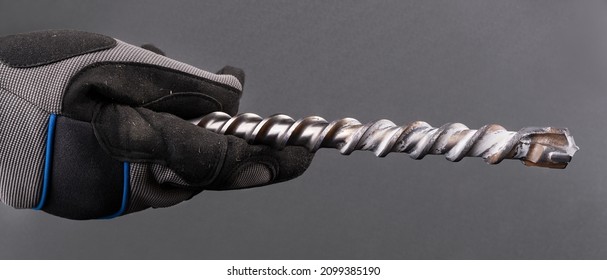 Hand with spiral fluted masonry drill bit with carbide tip on a gray background. Closeup of construction worker in protective glove holding metal tool for drilling concrete or hard building materials.