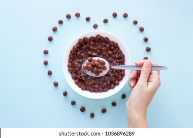 Hand with a spoon over a bowl with chocolate cereal balls on blue background.Dry breakfast cereal top view. Heart shape from flakes. Concept for a healthy diet.