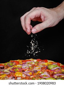 Hand sprinkling Parmesan cheese on pizza with cheese, bell pepper, red onions, bacon and pepperoni on dark background