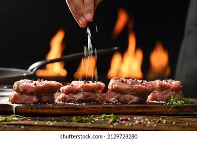 Hand sprinkling salt and seasoning on raw tenderloin steak meat beef on wooden chopping board on a wooden table prepared for cooking with flames in the background.