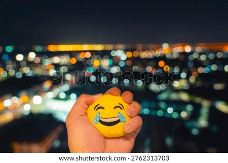 Hand squeezing crying smile face toy against blurred city night bokeh. Concept of depression, faking happiness, hidden sadness, mental health struggle, and stress management in modern society.