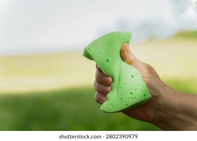 Hand squeezing a green cleaning sponge with a blurred nature background