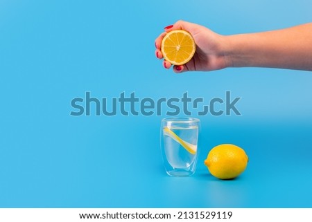 hand squeezing lemon juice in glass beaker with a slice of lemon and a lemon cut in half near it on an isolated blue background. Diet, vitamin c, weight loss concept