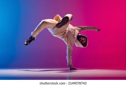 Hand stand. Studio shot of young flexible sportive man dancing breakdance in white outfit on gradient pink blue background. Concept of action, art, beauty, sport, youth. Dancer shows breakdance