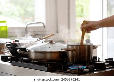 hand stirring a pot, kitchen full of pots cooking and cooking with gas, in the background a window with natural light and vegetation. - Powered by Shutterstock