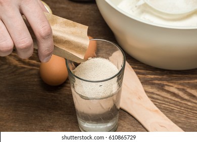 Hand Strewing Yeast From Packet Into A Glass Of Water. Preparing For Dough Making.