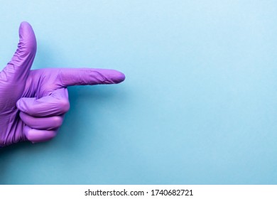 Hand Of Surgeon In Violet Medical Glove Showing Look Here Sign Gesture, Isolated On A Light Blue Background.