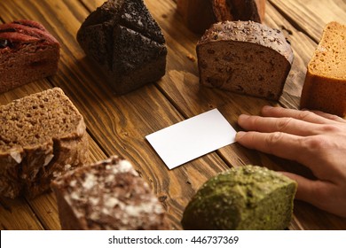 Hand takes blank business card of professional artisan baker presented in center of many mixed alternative baked exotic bread samples above wooden rustic table - Powered by Shutterstock