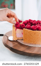 Hand takes slice of raspberry cake from wooden board with fresh berries in bright kitchen, dessert moment, homemade baking