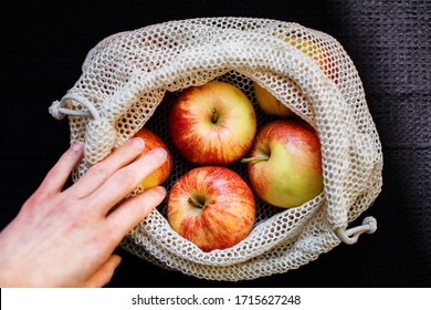 A Hand Is Taking An Apple From A Reusable Bag Full Of Apples. The Eco-friendly Bag Is Placed On A Black Tea Towel