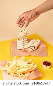 Man’s Hand Taking A Chicken Wrap On Napkin. Near Stands A Basket With French Fries On Yellow Table.                          