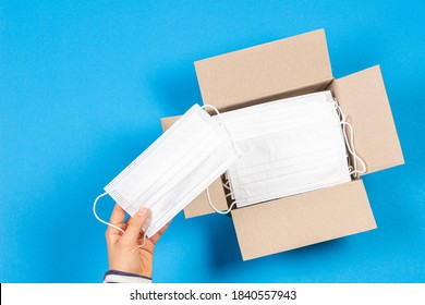 Hand Taking One Medical Mask From Open Paper Box With Lot Of Medical Masks On Light Blue Background. Top View