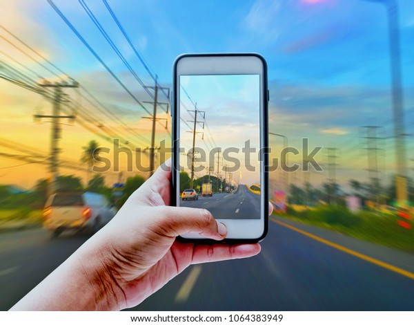 Hand taking a\
photo of city road in thailand and sunset light on moving car with\
smartphone | blurred\
background