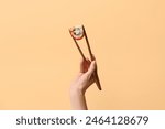 Hand with tasty sushi roll and chopsticks on beige background