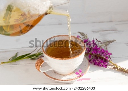 a hand from a teapot pours a drink (decoction) from the leaves and flowers of the medicinal plant ivan-tea (cypress, epilobium) in a mug. Traditional medicine, collection of useful herbs. Alternativ