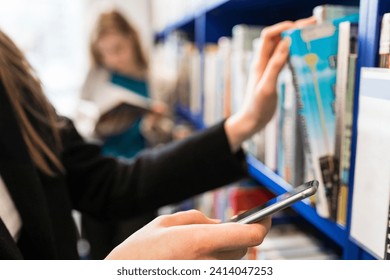 Hand of teenage girl holding cell phone while taking book from shelf in a public library - Powered by Shutterstock