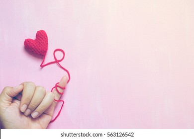 A hand that little finger bind with red yarn with crochet heart on painted pink canvas background - Shutterstock ID 563126542