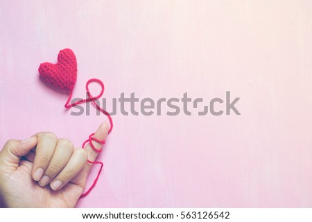 A hand that little finger bind with red yarn with crochet heart on painted pink canvas background