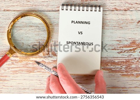 A hand thoughtfully presents a notebook displaying PLANNING VS SPONTANEOUS beside a golden magnifying glass and striped pencil on aged wood.