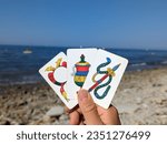 Hand of three Neapolitan cards: ace of Golds (asso di denari), ace of Cups (asso di coppe), ace of Swords (asso di spade). Used in Briscola or Scopa Italian card game. In the background: the sea.
