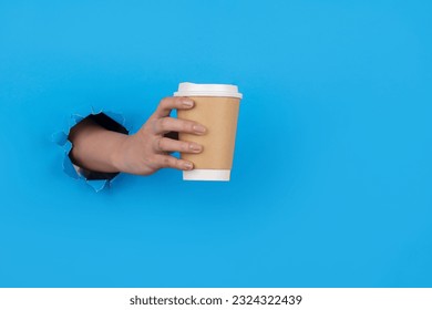 Hand through paper holding coffee cup on blue background