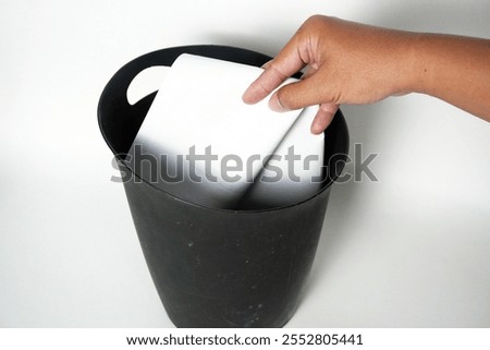 Hand throwing paper and tissue into a old minimalist trash can made of black plastic on a white background