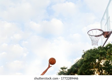 The Hand Throws The Ball Into The Basketball Hoop. Side View. Sky With Clouds In The Background. Concept Of Sports Games