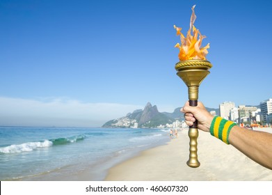 Hand Of Torchbearer Athlete Wearing Brazil Colors Sweatband Holding Sport Torch On Ipanema Beach With Two Brothers Mountain On The Skyline Of Rio De Janeiro, Brazil