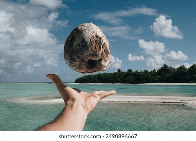 A hand tosses a coconut in the air against a backdrop of a tropical beach with clear turquoise waters, lush greenery, and a partly cloudy sky, capturing a moment of island fun and adventure. - Powered by Shutterstock
