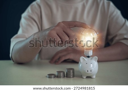 hand touching a glowing light bulb on a piggy bank with stacked coins, representing smart saving, investment growth, financial planning, innovation, business ideas, and long term wealth strategy.