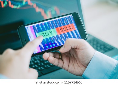 Hand Of Trader Holding Mobile Phone Touch Screen Showing Buy And Sell In Stock Market Order And Blurred Bakcground Of Laptop Show Financial Chart, Business Trading Concept