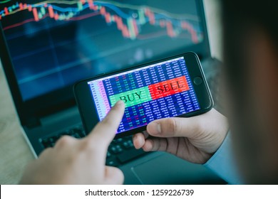 Hand Of Trader Holding Mobile Phone Touch Screen Showing Buy And Sell In Stock Market Order And Blurred Bakcground Of Laptop Show Financial Chart, Business Trading Concept