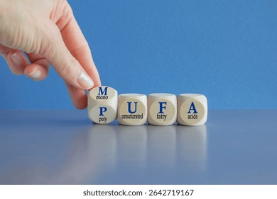 Hand turns cube and form the acronym MUFA (Monounsaturated Fatty Acids) and PUFA (Polyunsaturated Fatty Acids). Beautiful blue background, grey table. Business and medical concept. Copy space.